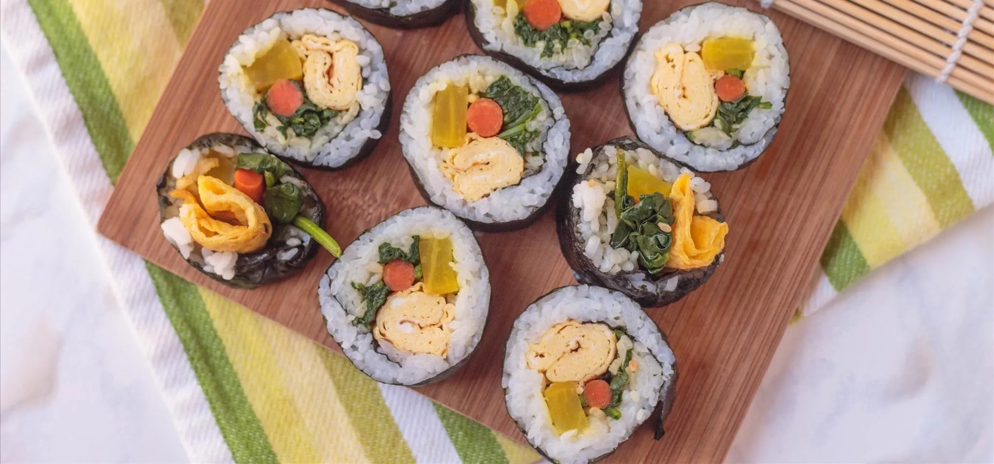 Colorful sushi rolls arranged on a wooden plate on top of a striped green and yellow tea towel.