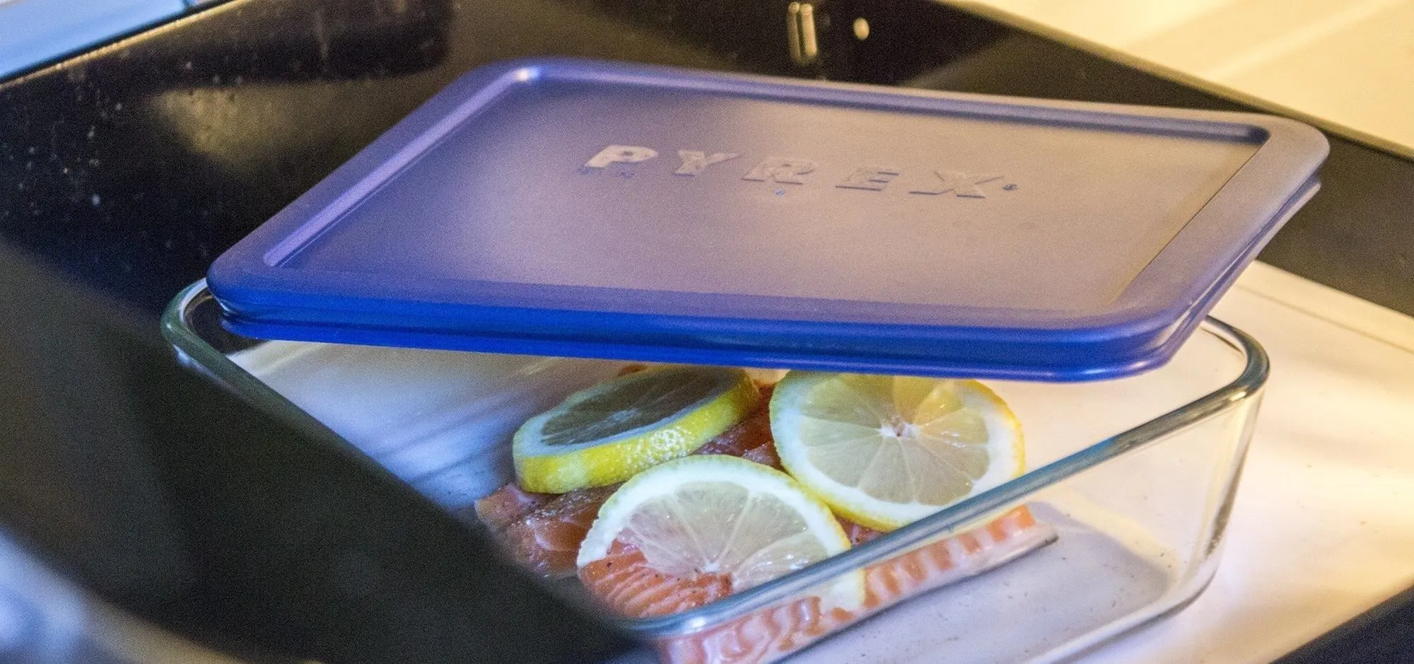 Salmon fillets with lemon slices covered by a blue lid in a glass dish about to be microwaved.