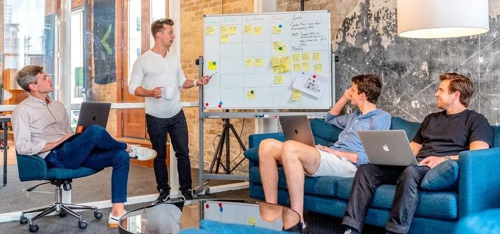 Business meeting in a modern office with a presenter discussing ideas on a whiteboard while colleagues listen and take notes.